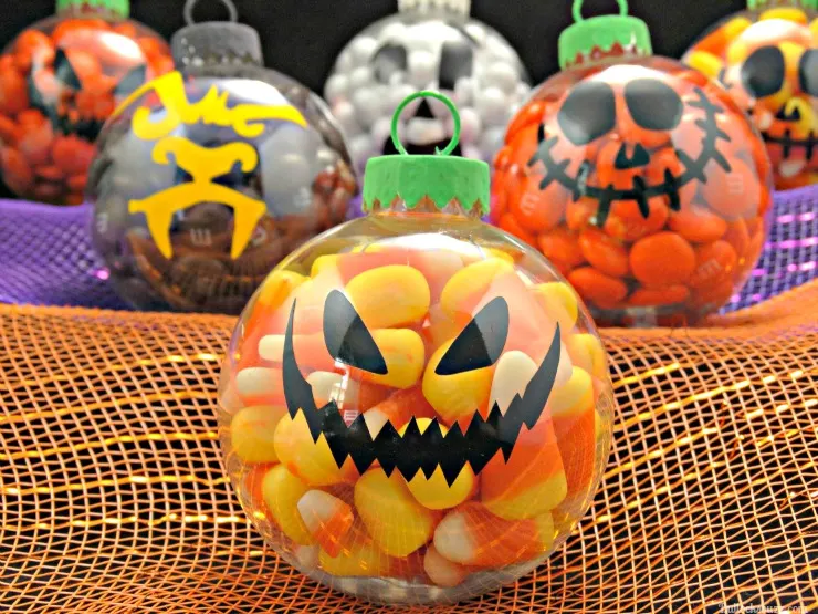 A candy corn filled pumpkin DIY Halloween Candy Ornament in front of several other candy-filled DIY Halloween ornaments displayed on orange and purple fabric.