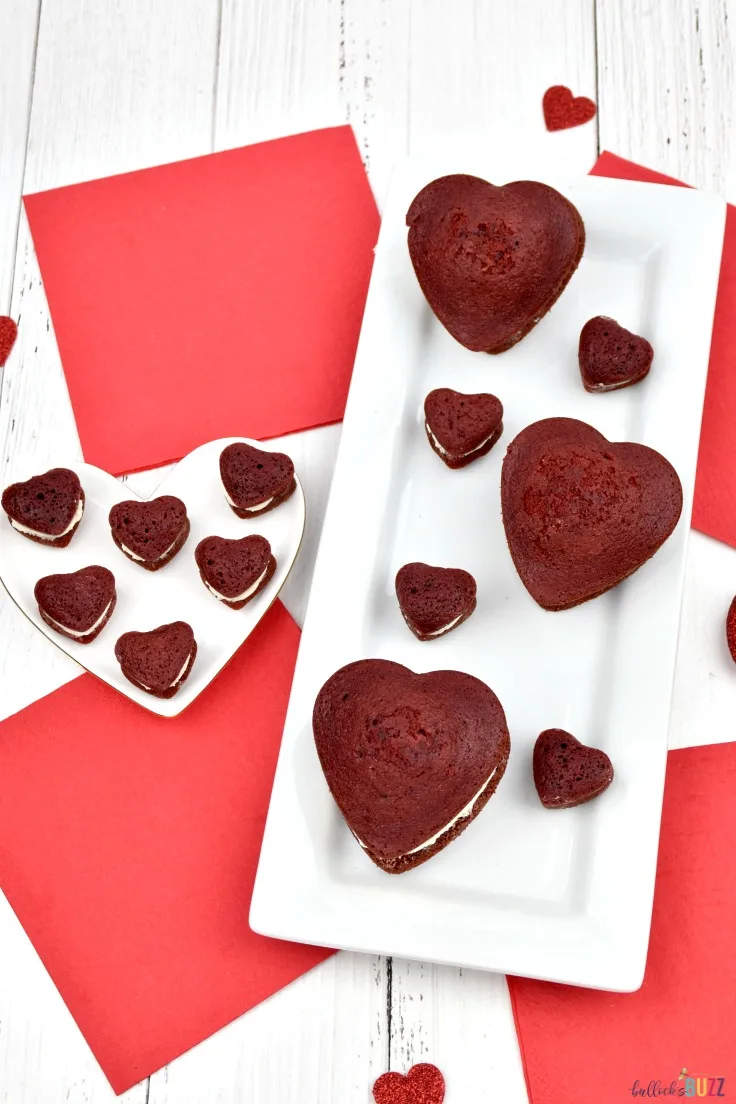 Large and miniature heart-shaped red velvet whoopie pies on white plates with a red and white background.