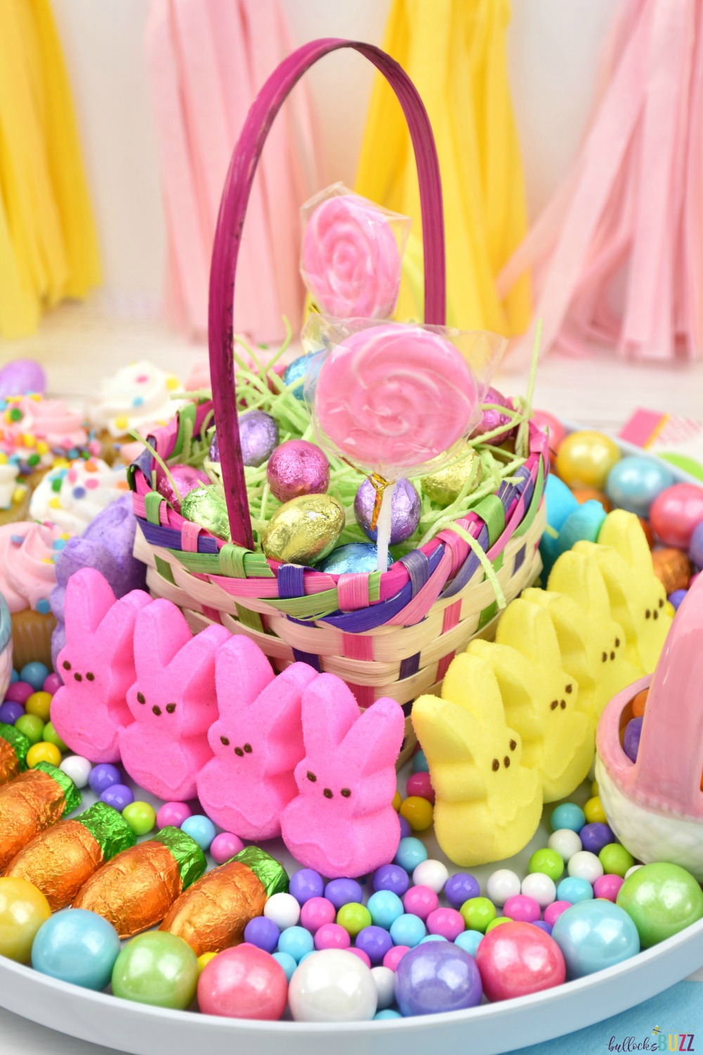 An Easter CAndy Board featuring a small Easter basket in the middle that is filled with edible green grass and chocolate Easter eggs surrounded by an assortment of pastel colored Easter candy and Easter treats.