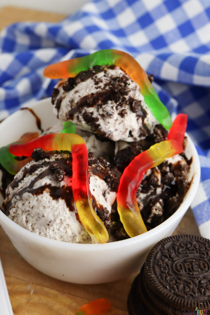 close up of ice cream sundae topped with crushed Oreo cookies, chocolate syrup, and gummy worms in a whie bowl
