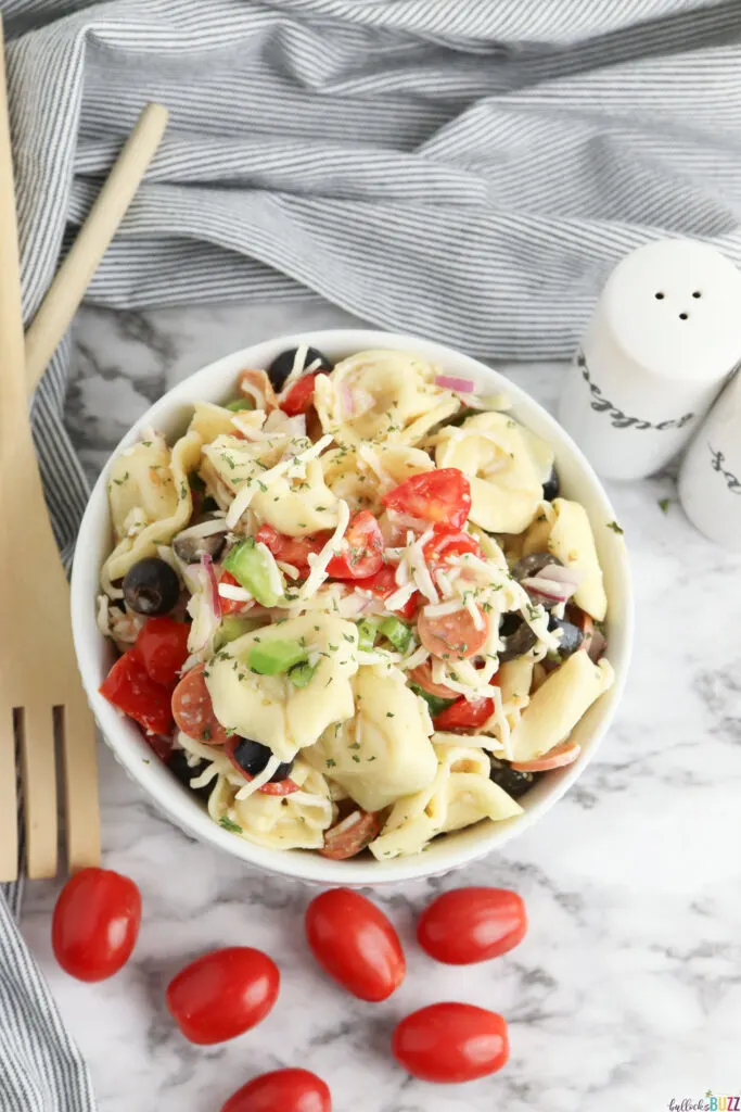overhead image of finished tortellini pasta salad in a bowl with tomatoes and wood cutlery next to it