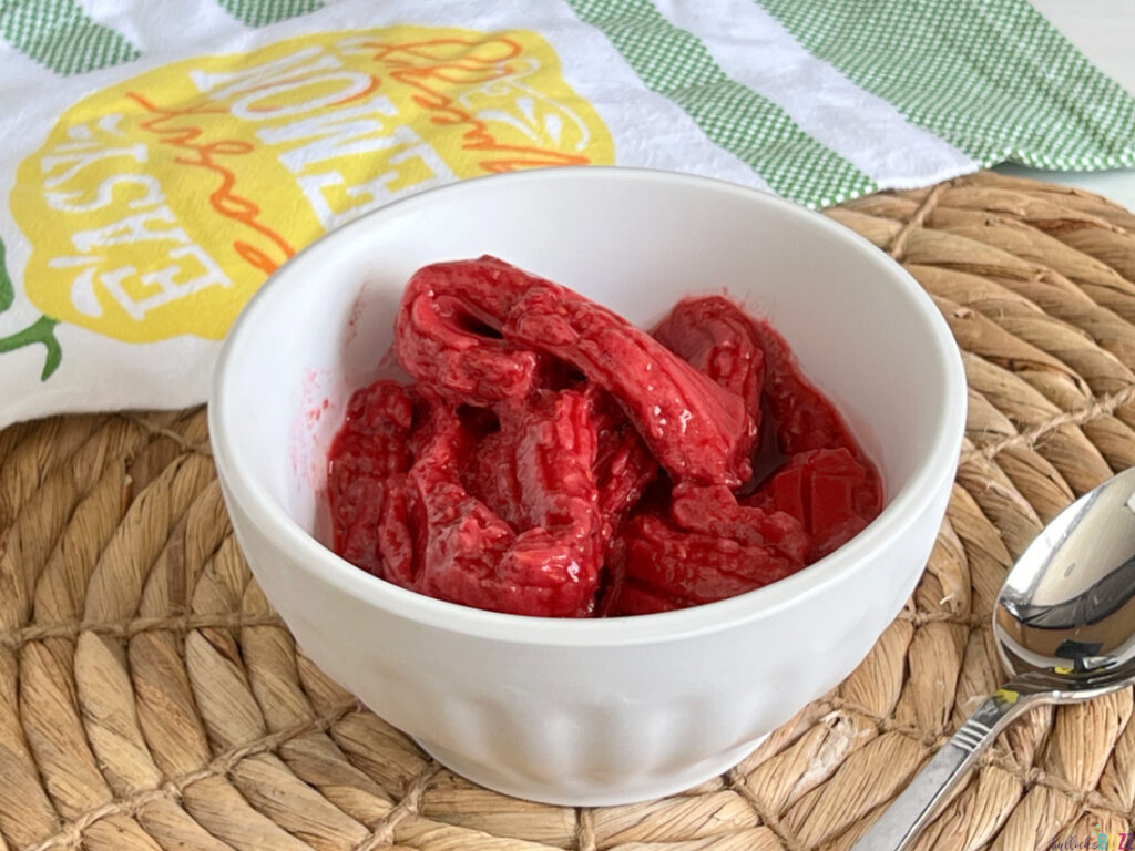 A white bowl full of homemade raspberry sorbet made in the Uber Appliance Sorbet Maker that is sitting on a rattan placemat with a spoon.