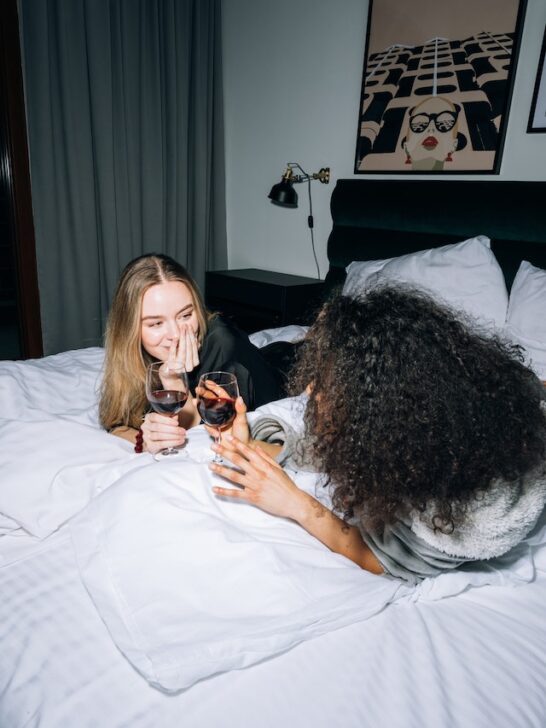 two friends talking while laying on bed drinking wine as a girls night in activities