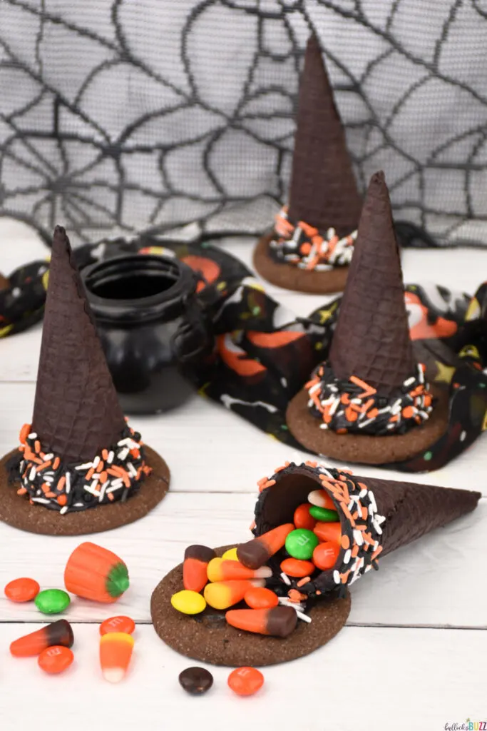 Unopened ice cream cone witch hats surrounding one open witch hat with its candy spilling out onto the table.