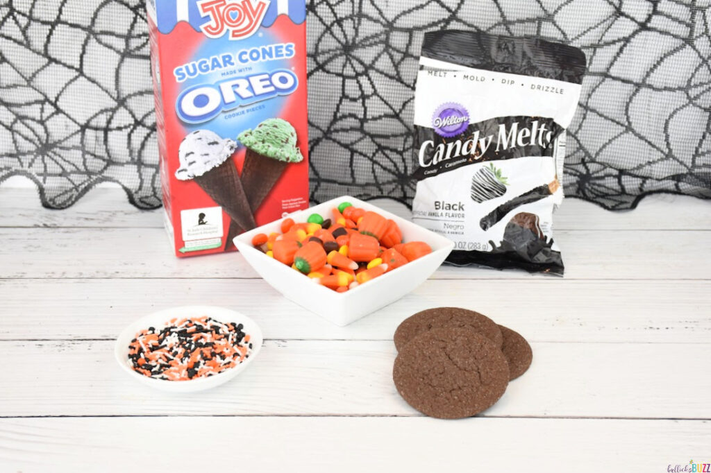 Ingredients you will need to make Halloween Candy-Filled Witch Hats including Oreo ice cream cones, black candy melts, Halloween sprinkles, fudge cookies and Halloween candy, laid out on a table.
