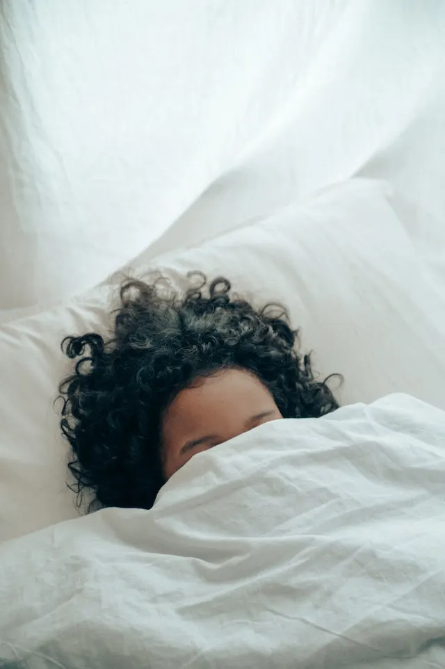 Woman with curly hair sleeping with her face covered by a sheet and her black curly hair spread around her head. Sleeping with dry hair will help avoid a bad hair