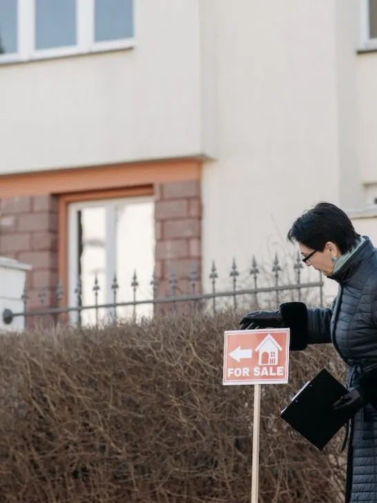 A woman posting a for sale sign in front of a home where the owner is selling a house as is for cash.
