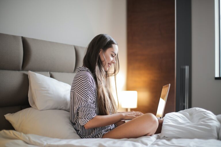 A woman sitting on her bed and smiling while typing on her laptop to create a fabulous dating profile