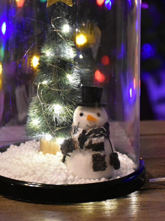 A close-up view of the adorable snowman inside the Christmas Cloche, positioned on a table with a beautifully lit Christmas tree in the background.