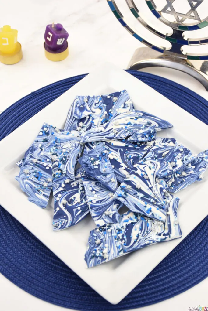 Finished Hanukkah Bark Candy on a white plate atop a blue placemat. accompanied by a festive menorah—a delightful holiday tableau of sweet celebrations.