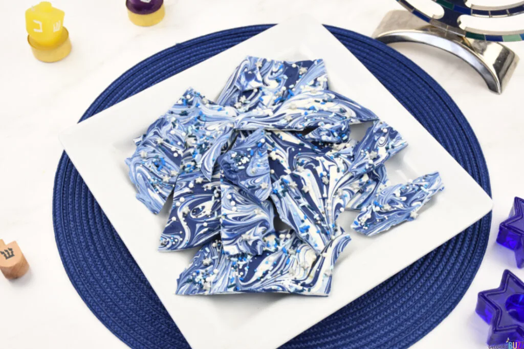 Hanukkah Candy Bark on a plate, set on a blue placemat with festive Hanukkah decor—a delightful and visually appealing holiday treat.