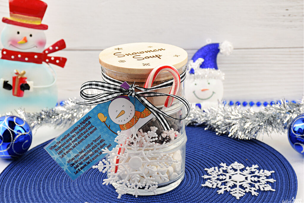 Side view of the completed Snowman Soup Hot Cocoa Jar, prominently displaying the enchanting Snowman Soup poem attached to the jar with a ribbon. In clear view are the decorative snowflake ornament and a candy cane, beautifully complementing the layered contents of cocoa mix, marshmallows, and chocolate-peppermint blend inside the jar.