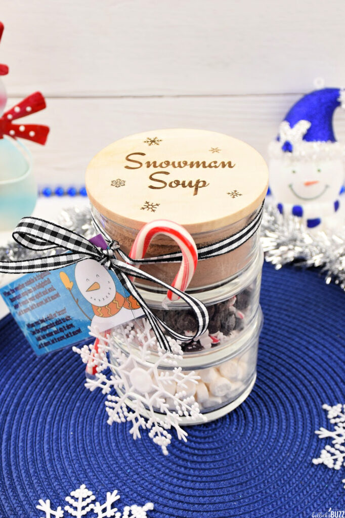 Image of the completed Snowman Soup Hot Cocoa Jar, showcasing its three distinct layers: rich cocoa mix at the bottom, fluffy mini marshmallows in the middle, and a festive blend of chocolate chips and peppermint on top. The jar is topped with an intricately engraved wooden lid, embellished with a decorative ribbon, a snowflake ornament, and a classic candy cane for a cozy holiday touch.