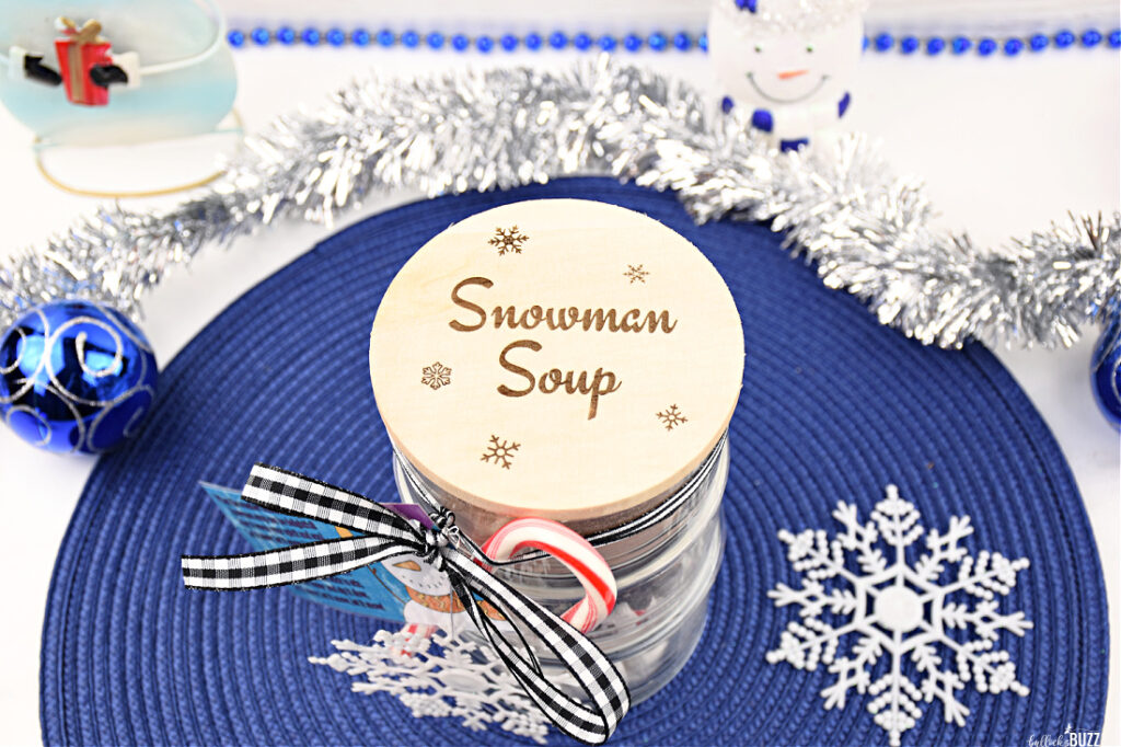 Overhead view of the completed Snowman Soup Hot Cocoa Jar, beautifully layered with homemade cocoa mix, marshmallows, and chocolate-peppermint topping, adorned with a laser-engraved lid, a festive ribbon, a snowflake ornament, and a candy cane.