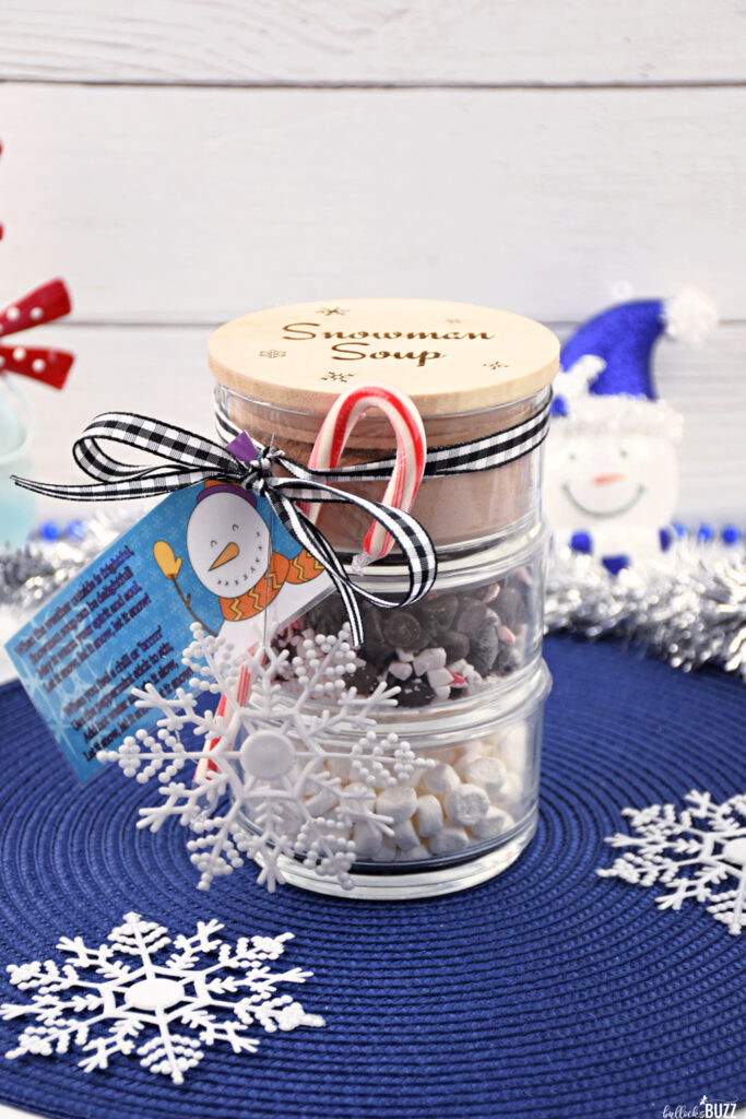 Final presentation of the Snowman Soup Hot Cocoa Jar, artfully composed with a laser-engraved lid featuring festive snowflake motifs. The jar layers include homemade cocoa mix, mini marshmallows, and a chocolate chips and peppermint mix, elegantly tied together with a ribbon. A candy cane and a snowflake ornament add the perfect finishing touches to this charming holiday craft."