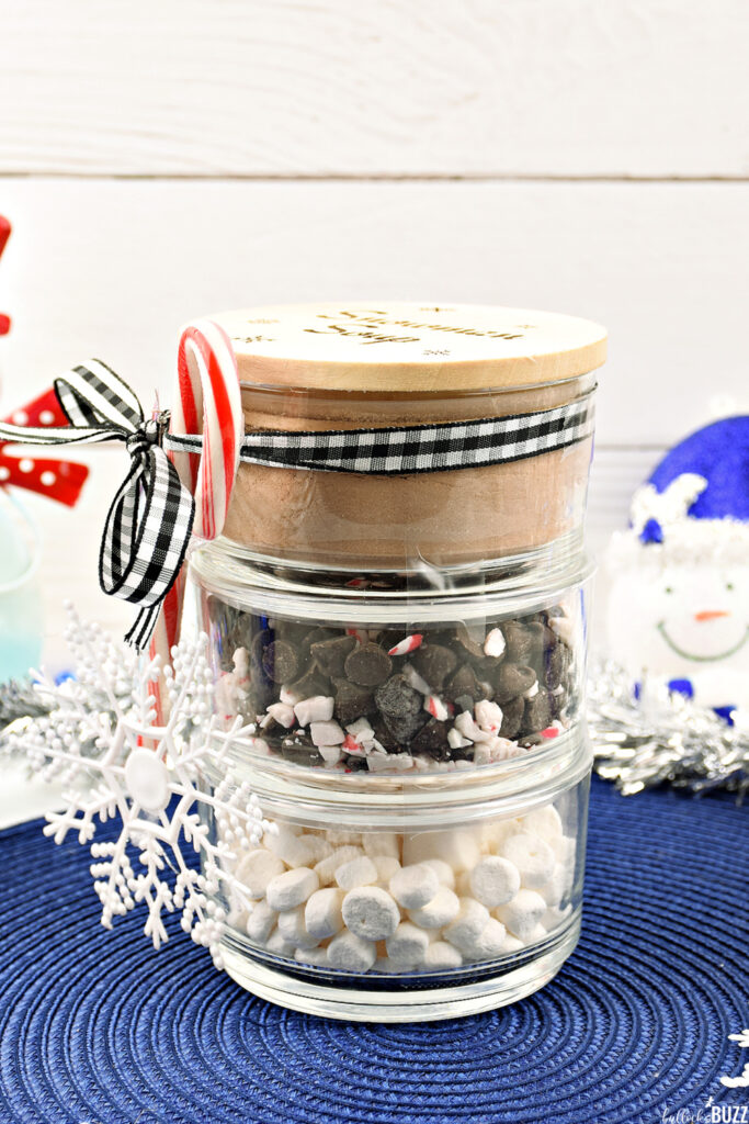 Image of the assembled Snowman Soup Hot Cocoa Jar, showing three stackable jars with the bottom layer filled with hot cocoa mix, the middle with mini marshmallows, and the top with chocolate chips and peppermint bits. The jar is completed with the engraved wooden lid on top.