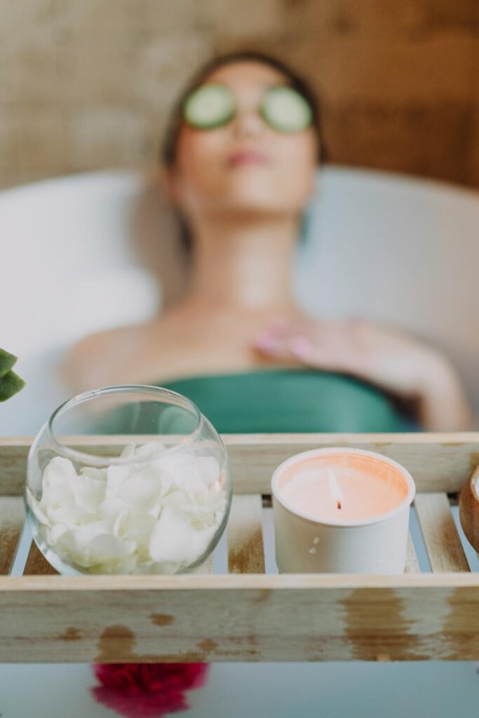A woman laying in a warm water tub with cucumber slices on her eyes. A tray with a candle and a vase of rose petals sits on a tray lying across the tub - all part of a relaxing spa day at home.
