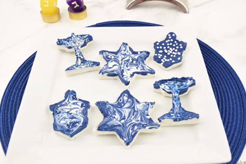 Six Pieces of Finished Hanukkah Candy in Star of David, Dreidel, and Menorah Shapes on White Plate Above Blue Placemat