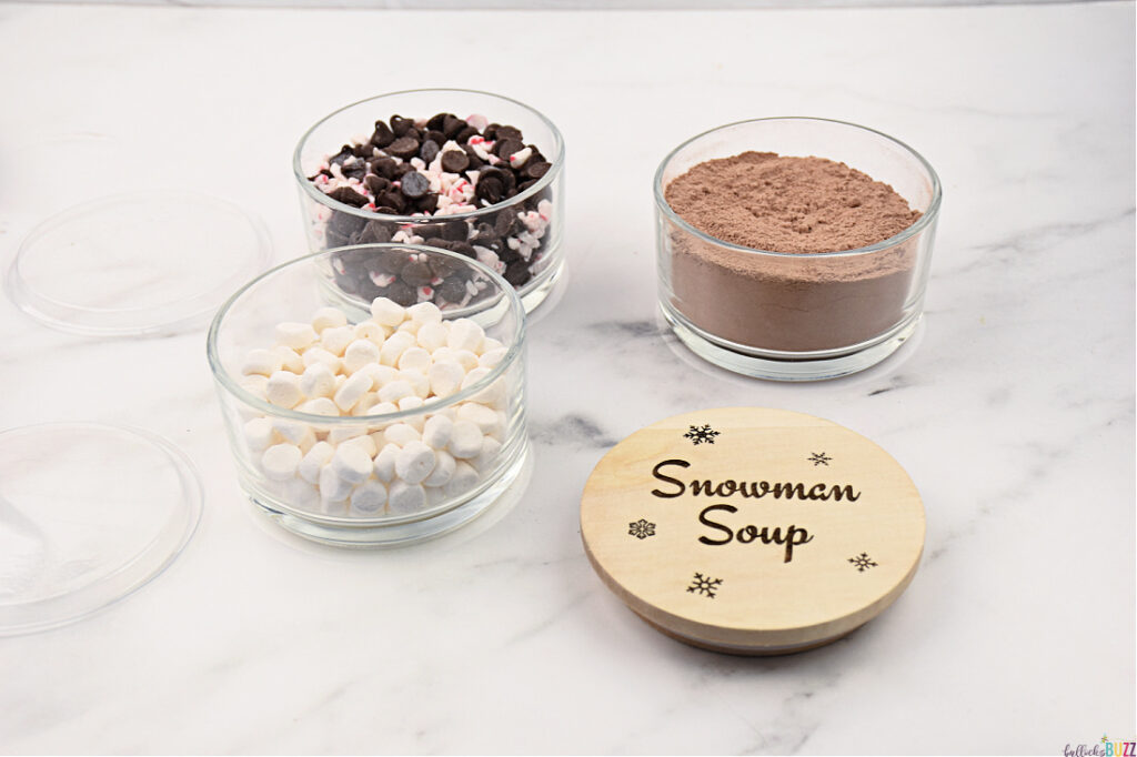 Three separate stackable jars arranged side by side, each filled with a different ingredient for Snowman Soup: one with homemade hot cocoa mix, another with fluffy mini marshmallows, and the third with a blend of chocolate chips and peppermint bits. In front of them lies the engraved wooden lid, showcasing the words 'Snowman Soup' and snowflake designs.