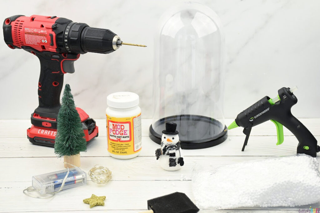 Flat lay of crafting supplies for DIY Christmas Cloche, including cloche, mini snowman, mini Christmas tree, styrofoam snow, hot glue gun, mini star, and LED lights, on a white background.