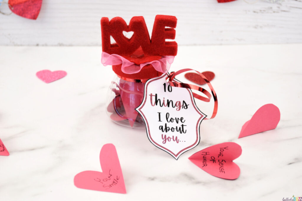 Red paper hearts with handwritten messages scattered around a jar filled with more cut-out hearts for '10 Things I Love About You' Valentine's gift.