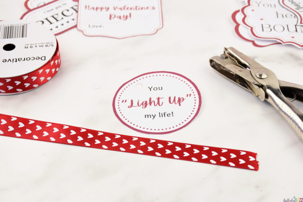 A cut out Valentine's tag with a hole punched in the corner, a spool of Valentine's ribbon, and a hole puncher on a table.