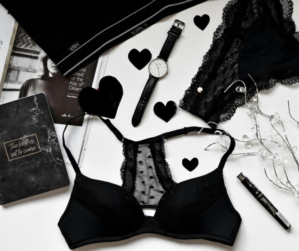 A black bra, black watch, mascara, magazine, diary, and black decorative hearts laid out on a white background