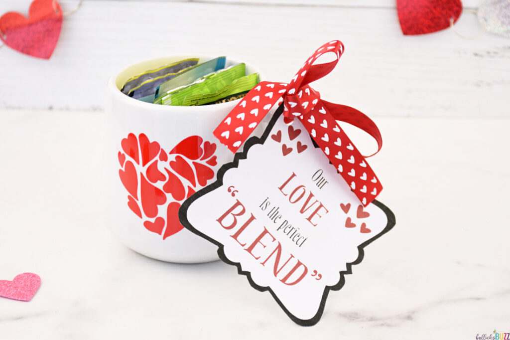 A white mug adorned with a red heart, filled with bags of tea, and featuring a 14 Days of Valentines tag that reads 'Our Love is the Perfect Blend'.