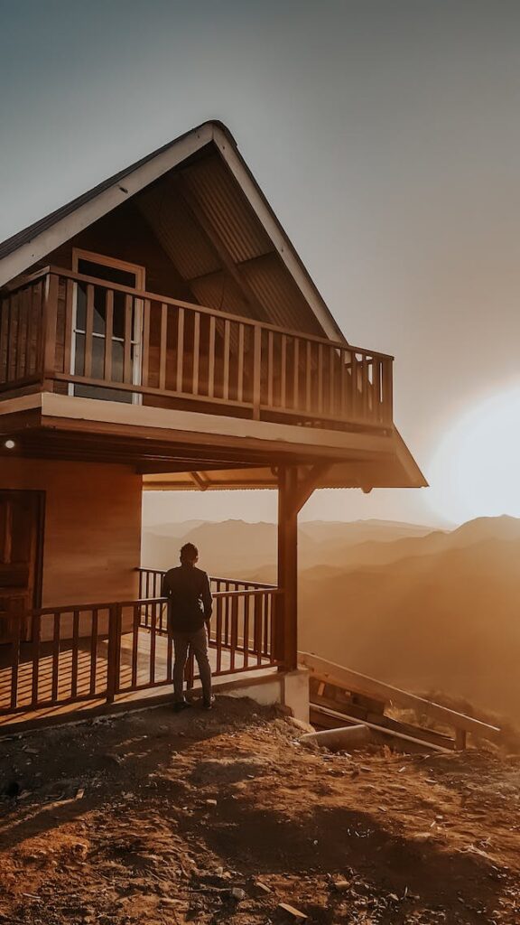Мужчина стоит на крыльце горной хижины и смотрит на горы.  Покупка коттеджа для отдыха – одна из самых популярных стратегий инвестирования в недвижимость.