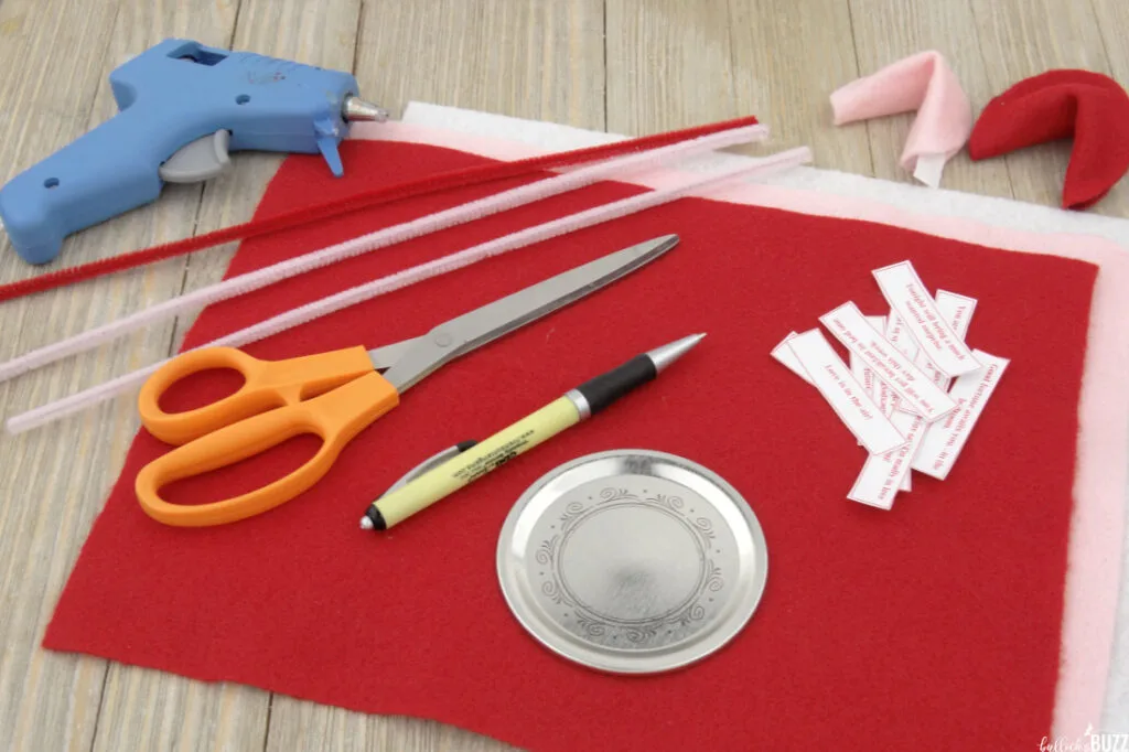 An image showing the supplies you will need to make DIY Felt Fortune Cookies for Valentines Day including felt, scissors, hot glue gun, glue sticks, mason jar lid, pen, and printable fortunes.