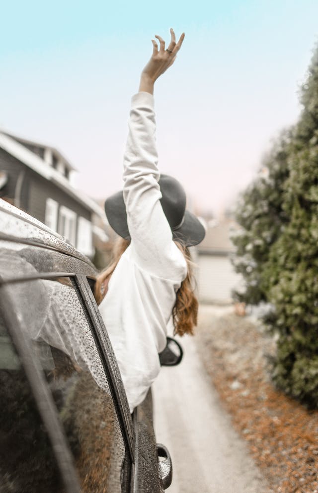 A girl wearing a hat and hanging out the window of a car heading off on a girls' road trip