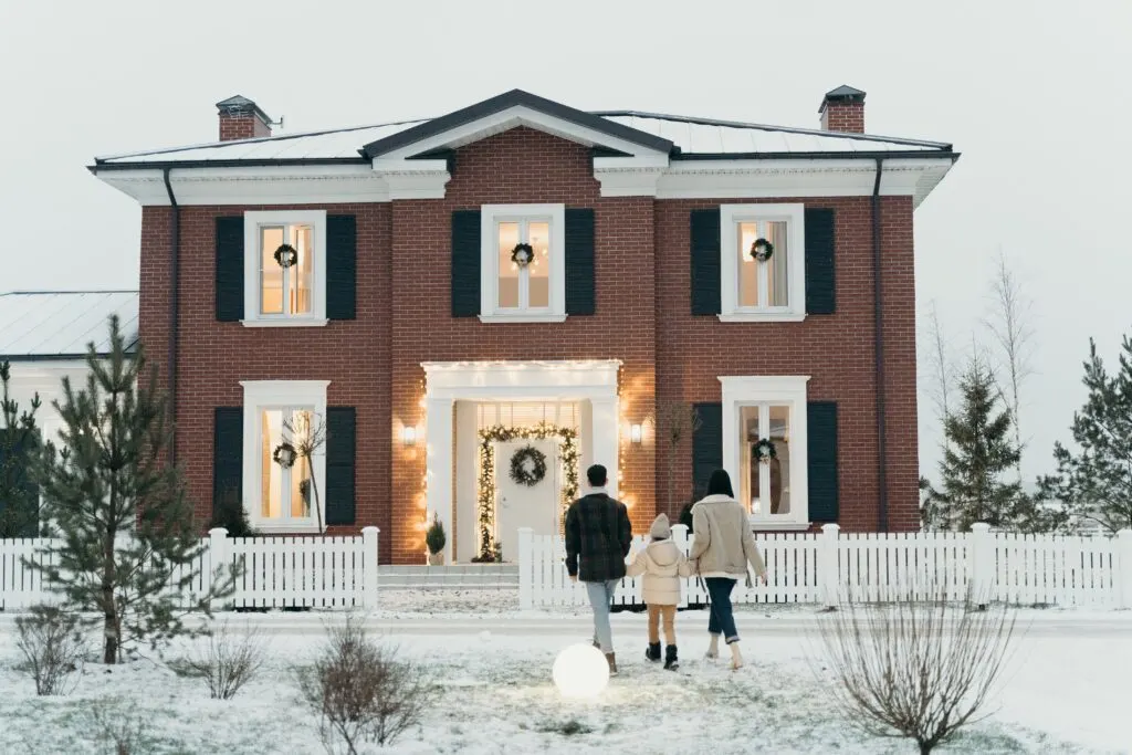 A family waling up to a pretty brick house decorated for the holidays with a snow-covered, healthy yard thanks to winter lawn care tips.