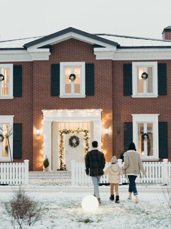 A family waling up to a pretty brick house decorated for the holidays with a snow-covered, healthy yard thanks to winter lawn care tips.