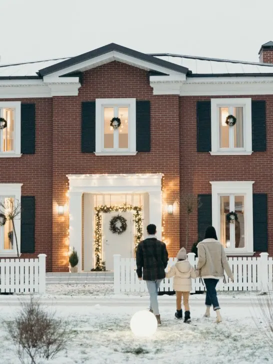 A family waling up to a pretty brick house decorated for the holidays with a snow-covered, healthy yard thanks to winter lawn care tips.