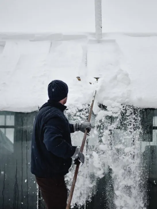 A man removing snow from his roof as part of winter roof maintenance tips.