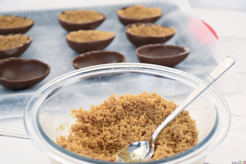 Close up of graham cracker mixture in a bowl with some of the hollow eggs filled sitting in the background