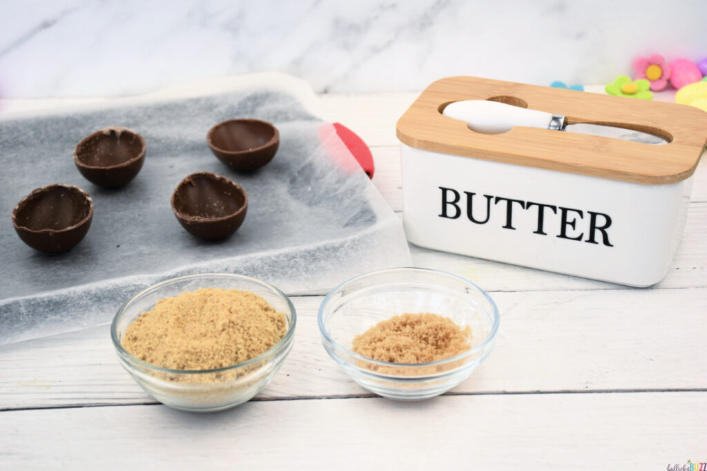 Ingredients for cheesecake crust in separate bowls: melted butter, crushed graham crackers, and brown sugar, ready for mixing.