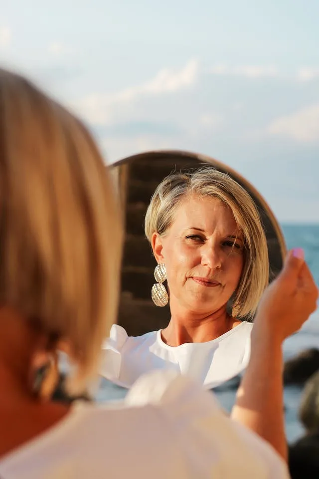 Beautiful middle age woman looking in the mirror at her skin. One major benefit of collagen induction therapy is that it helps keep skin youthful and healthy looking.