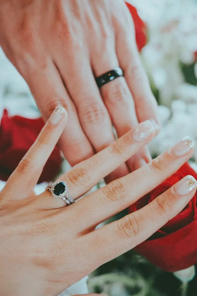 A woman's hand with an emerald and diamond engagement ring in front of  another hand with a tungsten ring