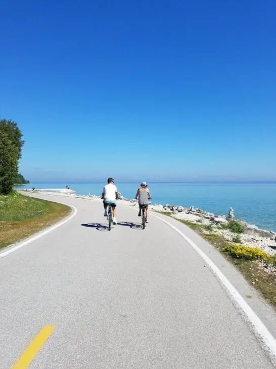 Two people riding bikes on a road by the shore. Riding bikes are an eco-friendly travel option for many reasons.