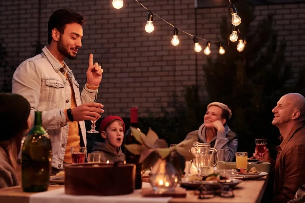 A man making a toast while people watch while sitting around a table under string lights at a backyard party