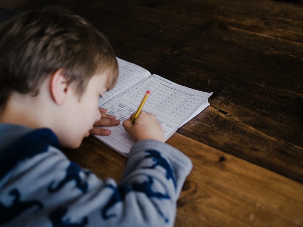 Маленький мальчик делает домашнее задание на коричневом столе.  Используйте эти стратегии, чтобы сделать домашнее задание менее пугающим для ваших детей.