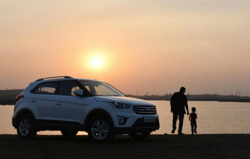 A man walking with his little boy by the lake at sunset with their car parked near them.