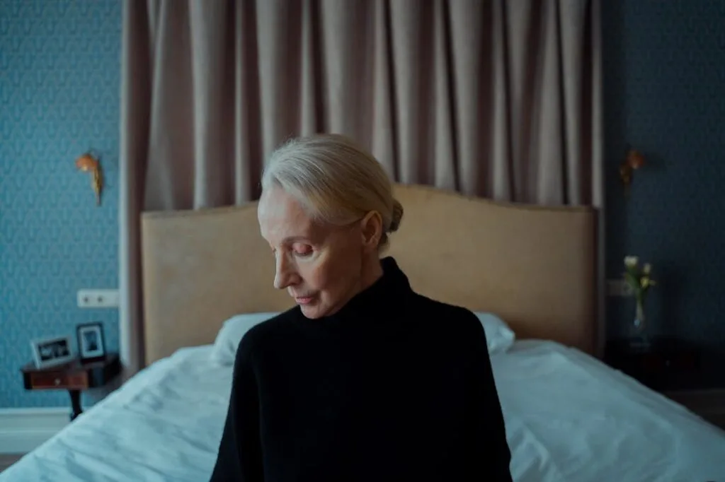 An elderly woman sitting on a bed dressed in black remembering