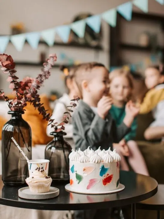 A birthday cake with kids playing in the background. Ensuring you have a large enough cake is an essential part of planning a child's birthday party for large groups.