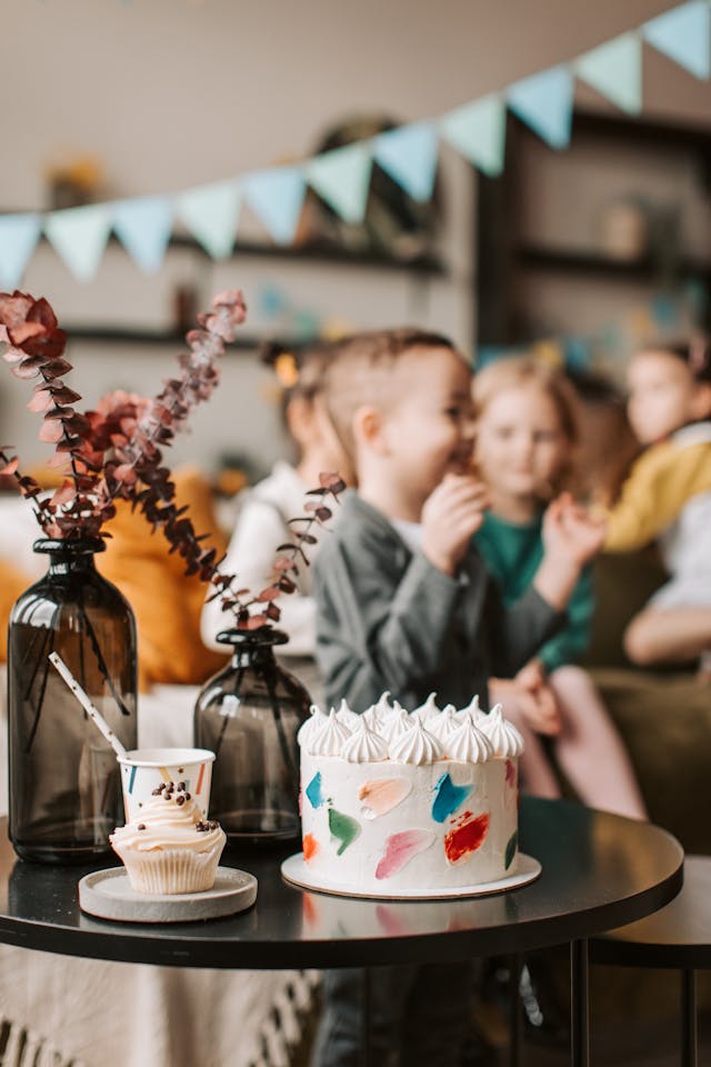 A birthday cake with kids playing in the background. Ensuring you have a large enough cake is an essential part of planning a child's birthday party for large groups.