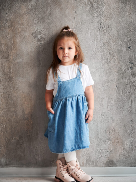 A little girl wearing a denim dress over a white t-shirt, one of twelve adorable summer dresses for your daughter