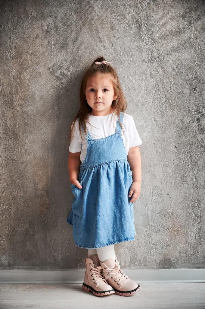 A little girl wearing a denim dress over a white t-shirt, one of twelve adorable summer dresses for your daughter