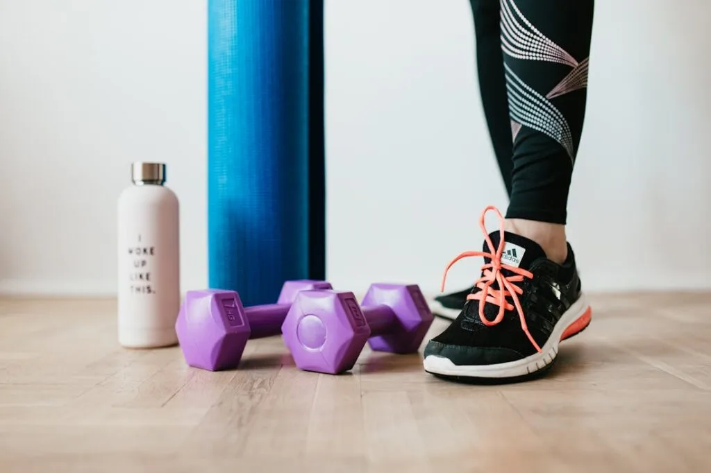 A yoga mat, hand weights, and a water bottle next to tennis shoes. These items are all great additions for when you are setting up your home gym.