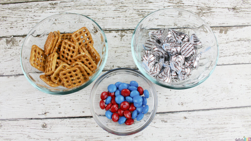 Pretzels, Hershey Hugs candy, and red and blue M&M's in small glass bowls as ingredients to make Patriotic Pretzel Hugs